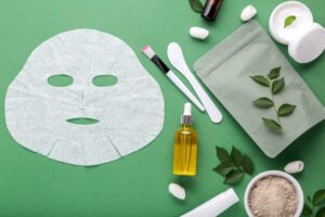 Fabric cosmetic face mask with set of cosmetics clay mask serum oil spatula brush, moisturizing cream in jar. Beauty spa treatments for facial skin care, cosmetology on green background with leaves